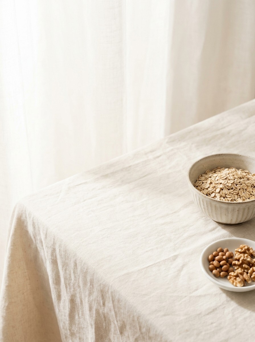 A bowl of rolled oats and a small white dish of soybeans and walnuts pushed to the lower-right of cream linen, the upper-left left empty.