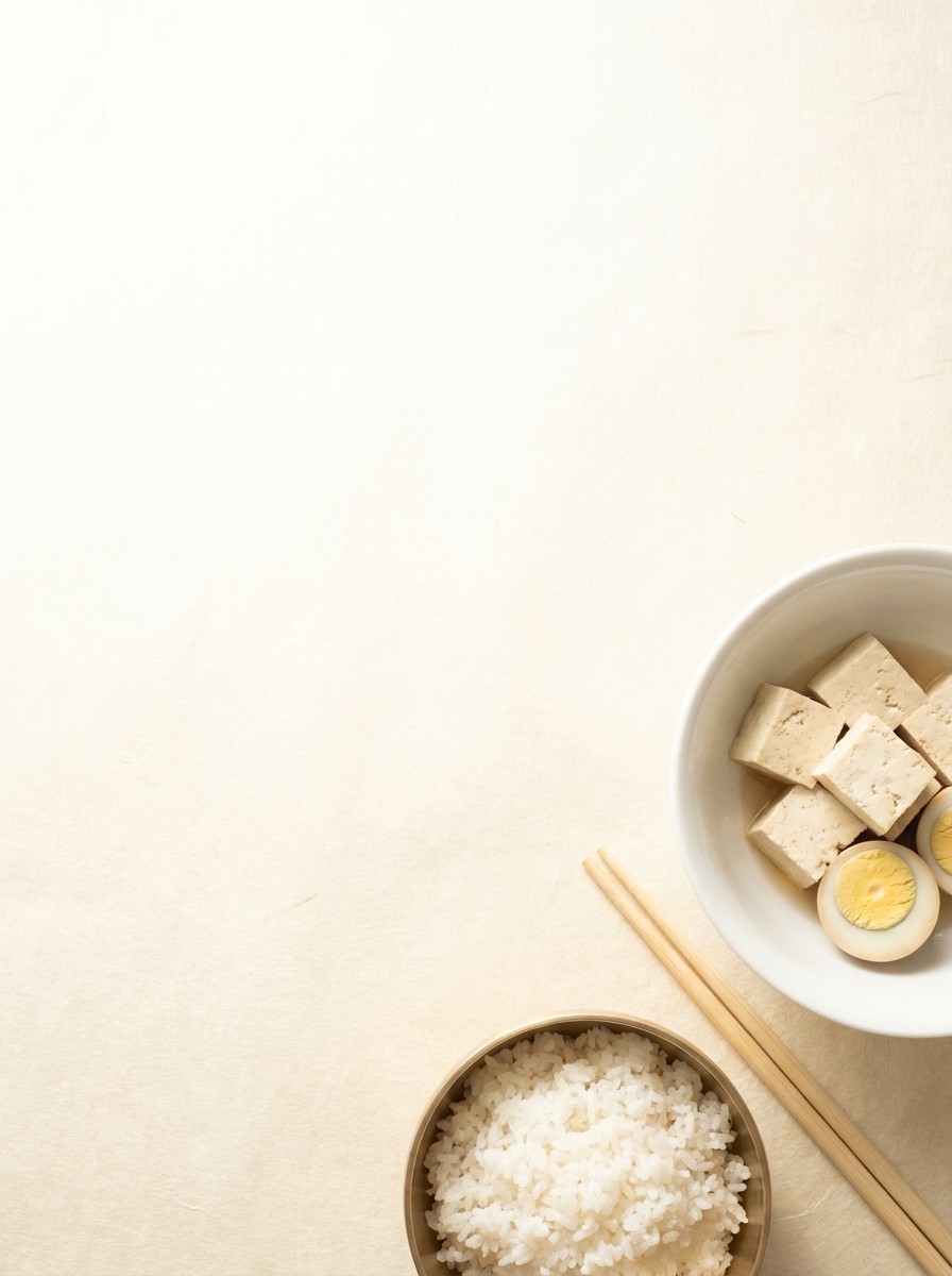 A white bowl of tofu and boiled eggs, a brass bowl of rice and wooden chopsticks pushed to the lower-right of hanji paper, the upper-left left empty.