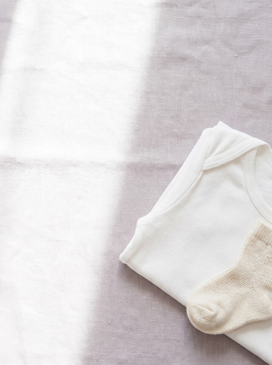 A folded white cotton garment and a cream knitted baby sock on pale lavender-grey linen, with open space at upper left.
