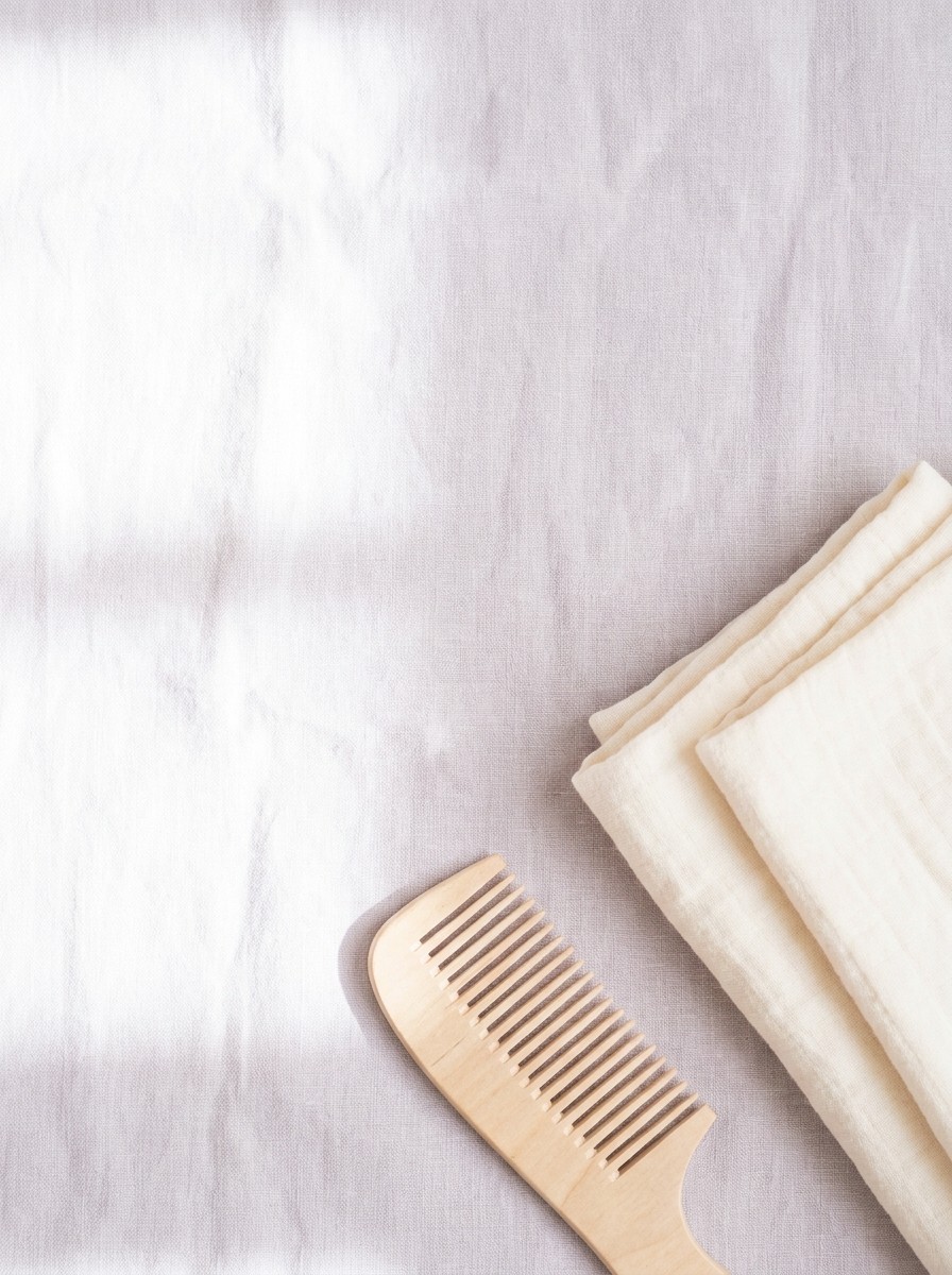 A wooden wide-tooth comb and a folded cream muslin blanket on pale lavender-grey linen, with open space at upper left.
