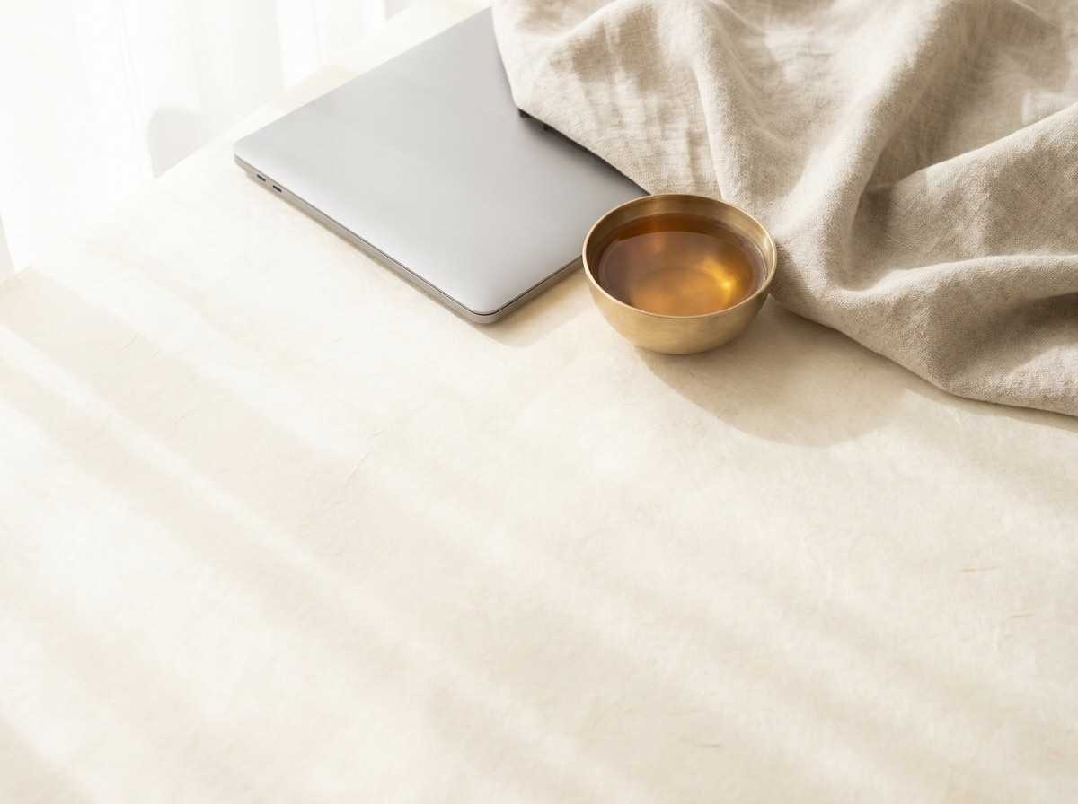 The corner of a closed silver laptop, a small brass bowl of warm barley tea and a folded oatmeal linen blanket on cream hanji in soft window light.