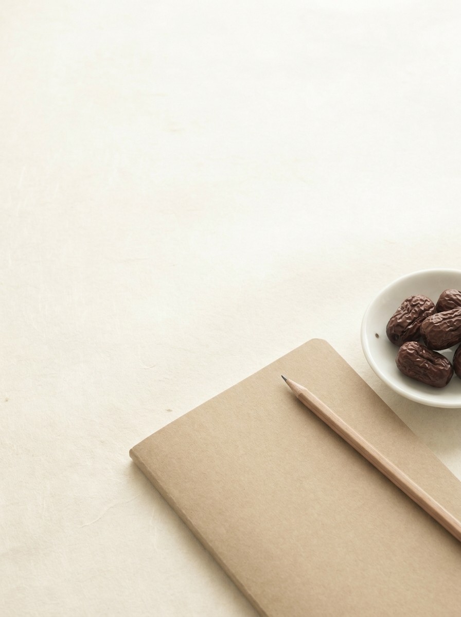 A closed kraft notebook with a wooden pencil and a white porcelain dish of dried jujubes pushed to the lower-right of cream hanji, the upper-left left empty.