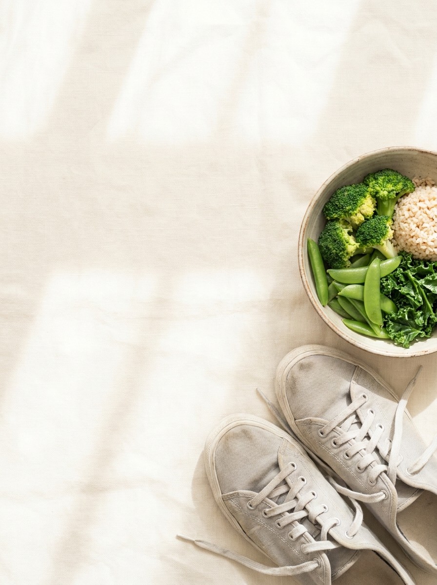 A pair of light gray walking sneakers and a bowl of steamed green vegetables with brown rice at the lower-right of cream linen, the upper-left left empty.