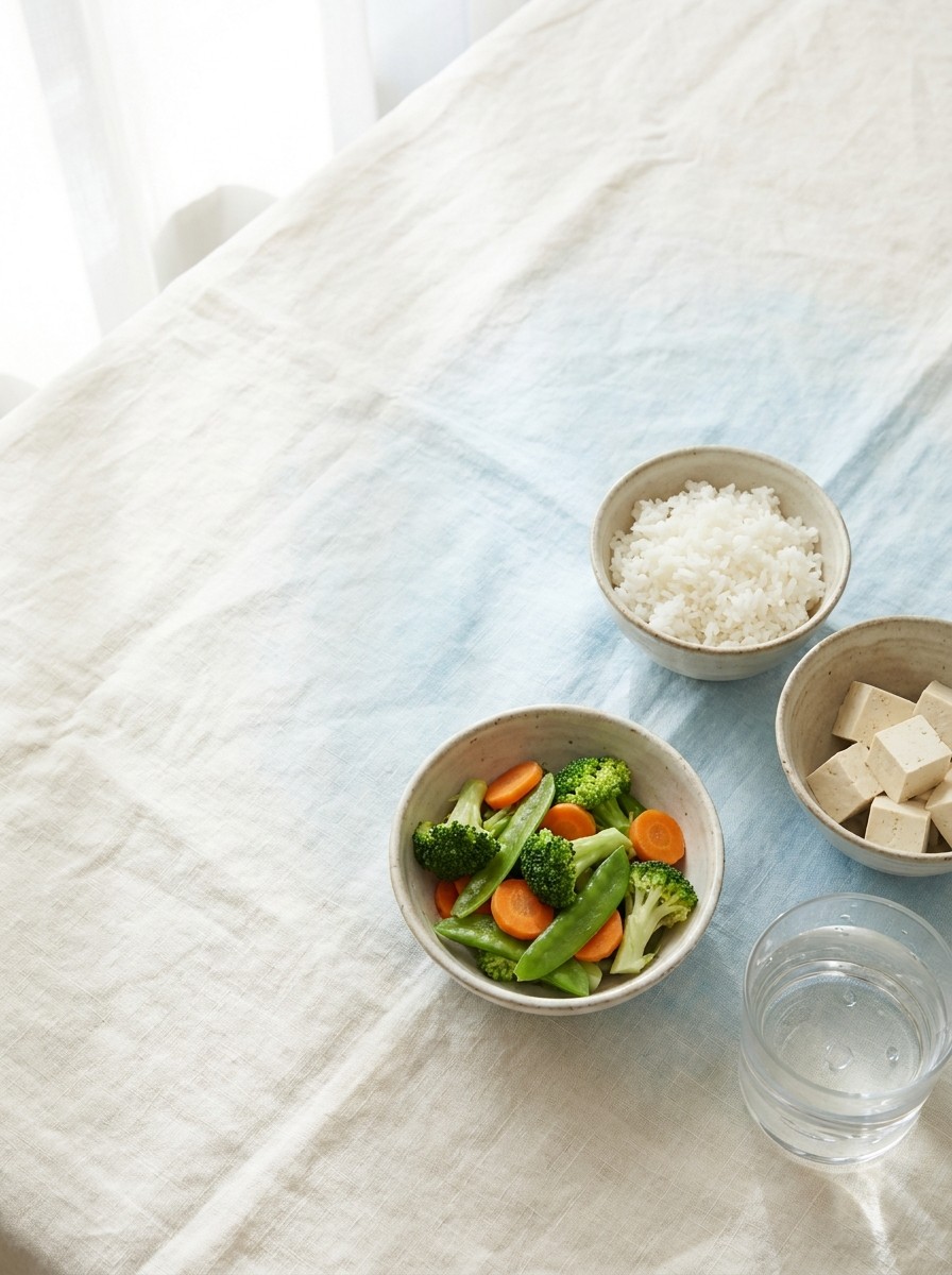 Three small ceramic bowls of rice, steamed vegetables and tofu with a glass of water on cream linen, with open space at upper left.