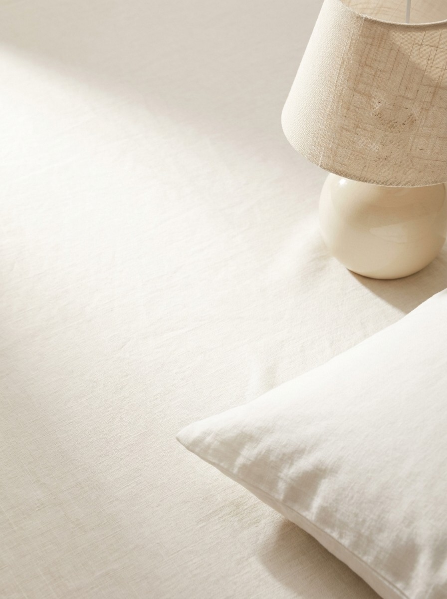 The corner of a white linen pillow and a switched-off ceramic bedside lamp pushed to the lower-right of cream linen, the upper-left left empty in soft morning light.