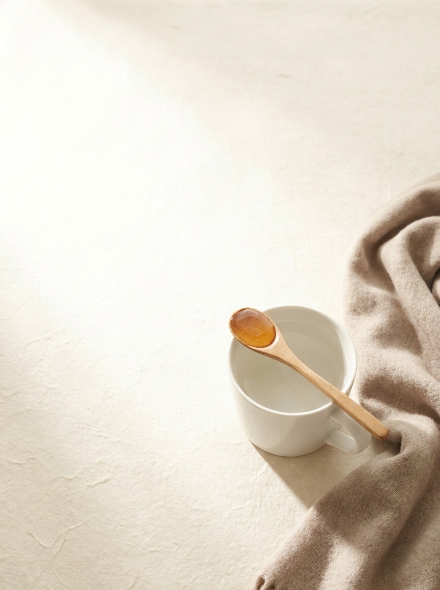 A white porcelain mug of warm water with a spoon of honey on its rim and a folded beige wool scarf at the lower-right of cream hanji paper, with bright empty space at the upper-left.