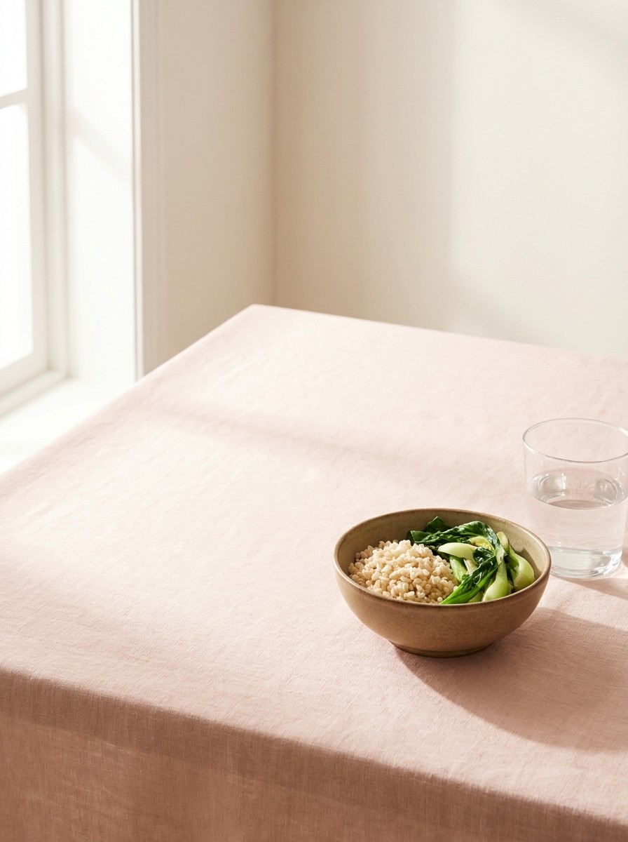 A small ceramic bowl of brown rice with steamed green vegetables and a glass of water on blush linen, with open space at upper left.