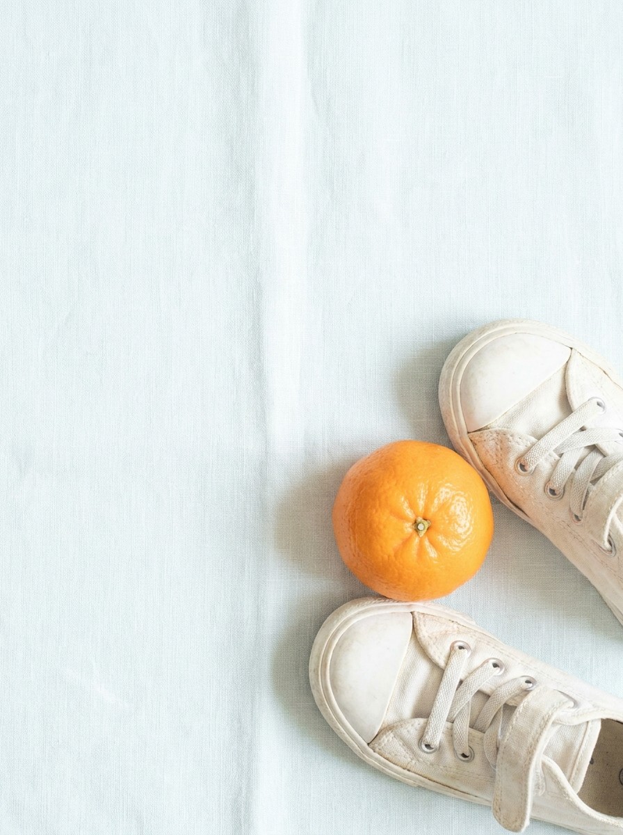 A pair of small white canvas child's sneakers and a mandarin orange on light cream linen with a pale sky-blue tint, with open space at upper left.