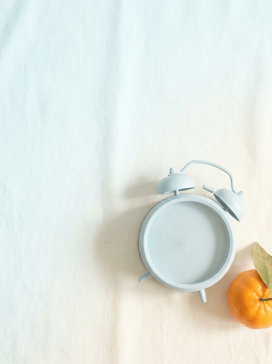 A small pale-blue alarm clock with a blank face and a mandarin orange on light cream linen with a pale sky-blue tint, with open space at upper left.