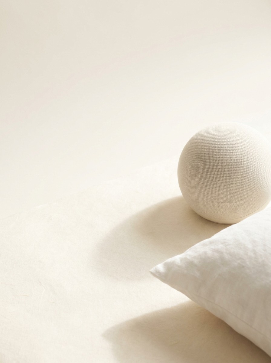 A small round cream fabric speaker and the corner of a white linen pillow at the lower-right of cream hanji paper, the upper-left left bright and empty.