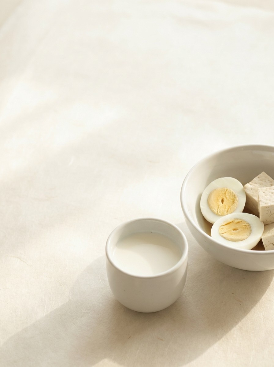 A white porcelain bowl of boiled eggs and tofu with a small cup of milk pushed to the lower-right of a hanji surface, the upper-left left empty.
