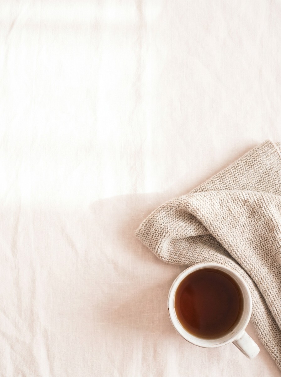 A soft folded oatmeal blanket and a white cup of warm barley tea on light cream linen, with open space at upper left.