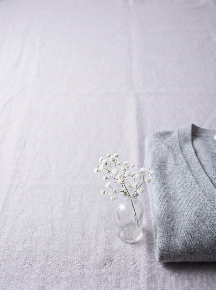 A sprig of white baby's breath in a small glass bottle beside a folded grey cardigan on lavender-grey linen, with open space at upper left.