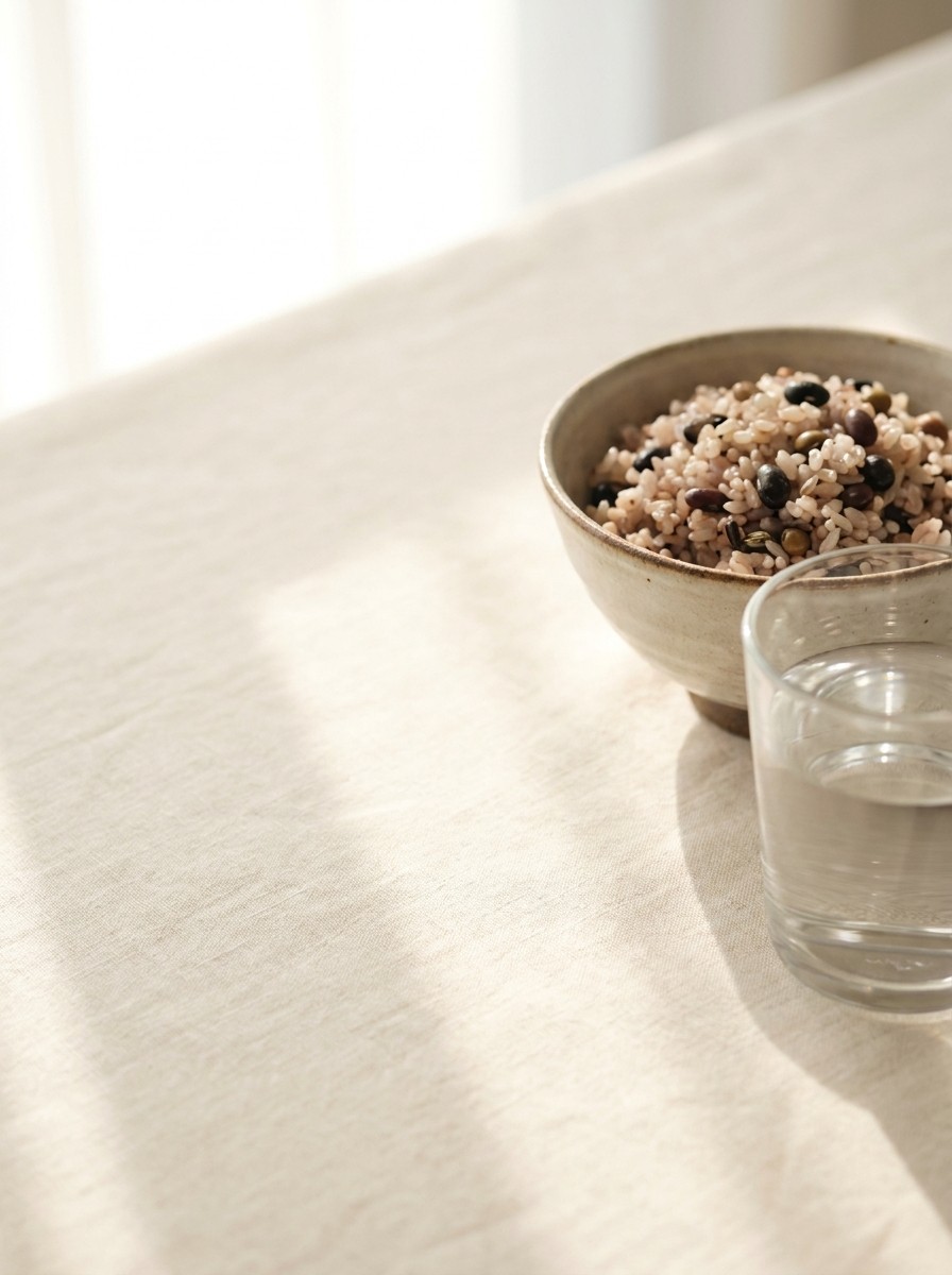 A glass of plain water and a bowl of mixed-grain rice with beans pushed to the lower-right of cream linen, the upper-left left empty in soft window light.