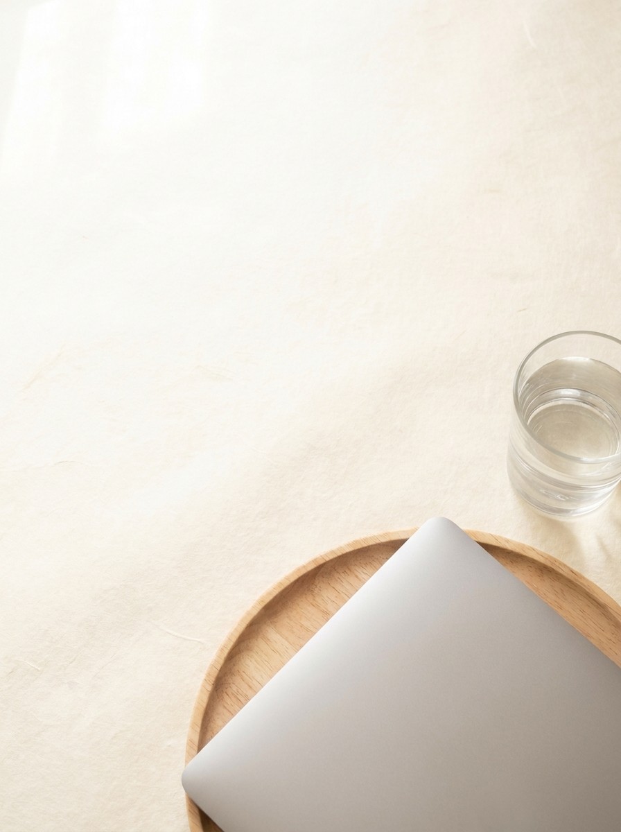 A closed silver laptop on a round wooden tray and a glass of cool water pushed to the lower-right of hanji paper, the upper-left left empty.