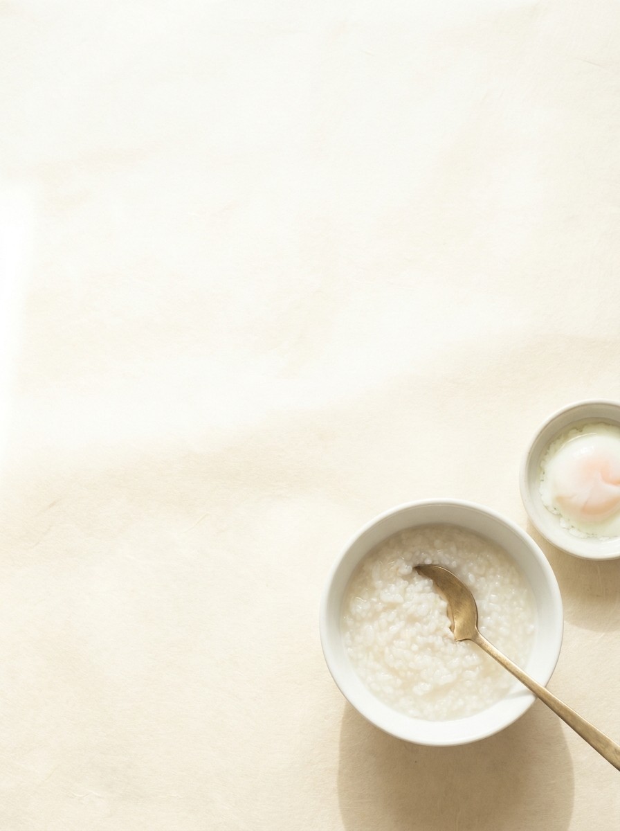 A small porcelain bowl of rice porridge with a brass spoon and a dish of soft egg pushed to the lower-right of hanji paper, the upper-left left empty.