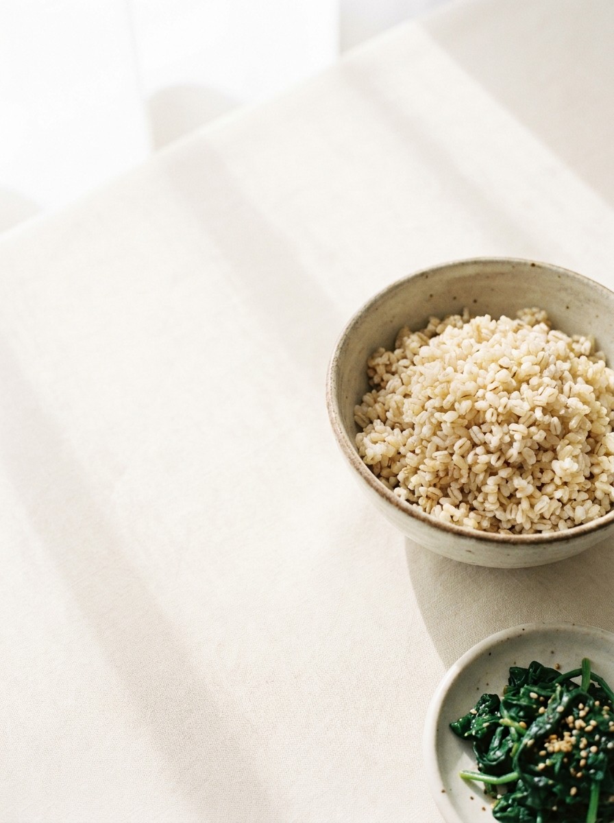 A bowl of brown rice with barley and a dish of seasoned spinach pushed to the lower-right of cream linen, the upper-left left empty.