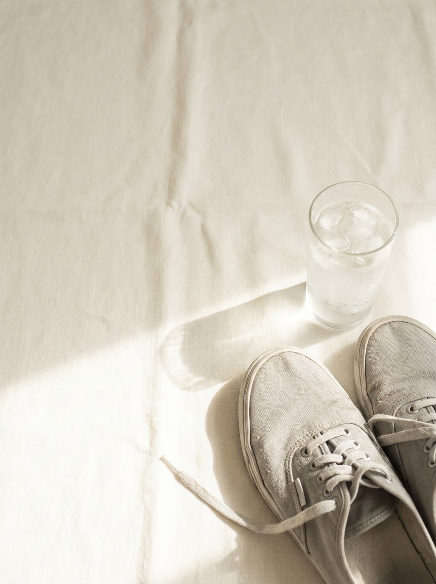 A pair of light gray canvas shoes and a tall glass of iced water pushed to the lower-right of cream linen, the upper-left left empty.