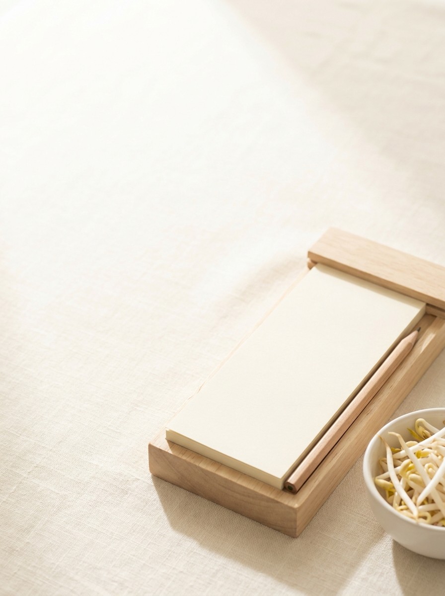 A blank desk calendar block with a pencil and a small white bowl of bean sprouts pushed to the lower-right of cream linen, the upper-left left empty in soft window light.