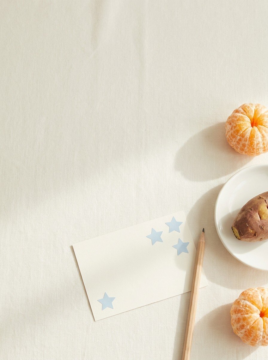 A blank card with pale blue star stickers, a pencil, a steamed sweet potato on a white saucer and tangerines on cream linen, upper left of the frame left empty