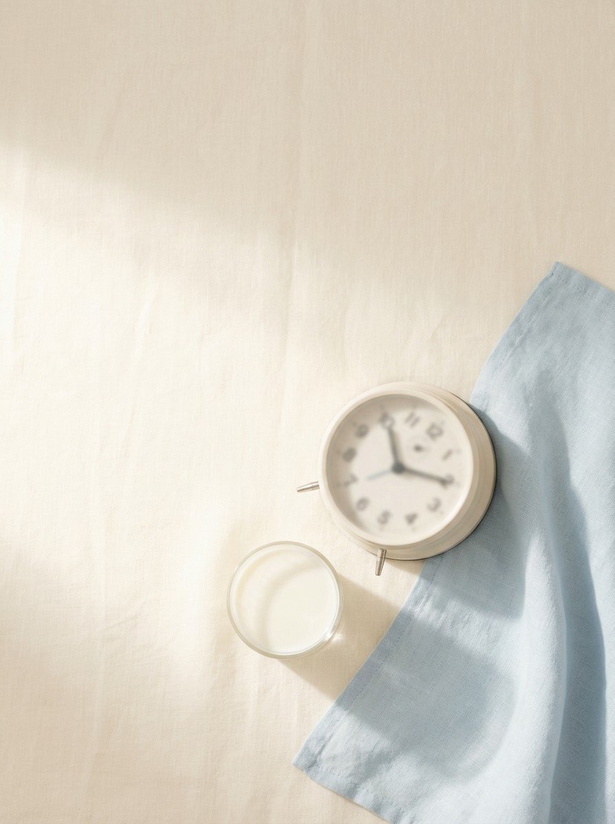 A small alarm clock lying on its side, the corner of a pale blue blanket and a glass of milk on cream linen, upper left of the frame left empty