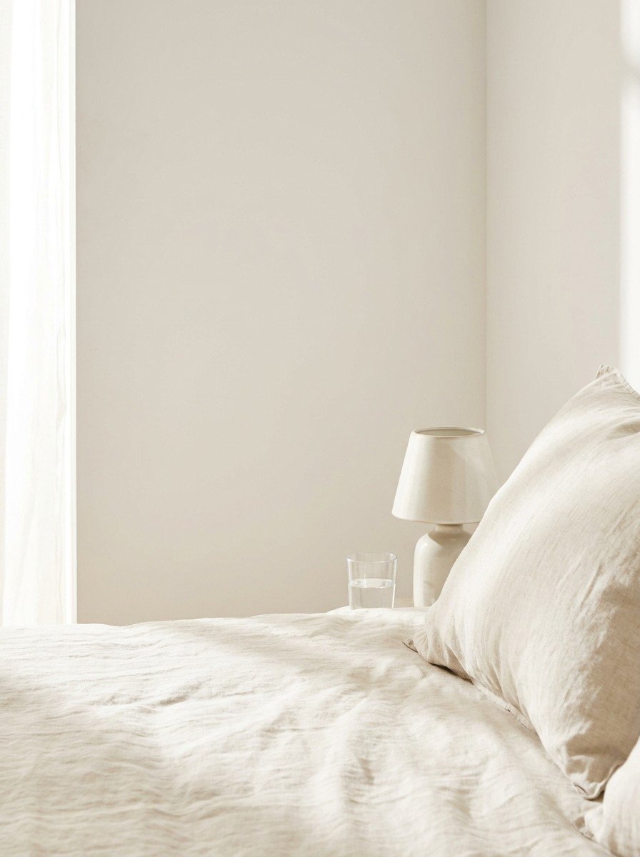 A linen pillow, a switched-off ceramic bedside lamp and a glass of water pushed to the lower-right, with an empty pale wall on the upper-left in soft morning light.