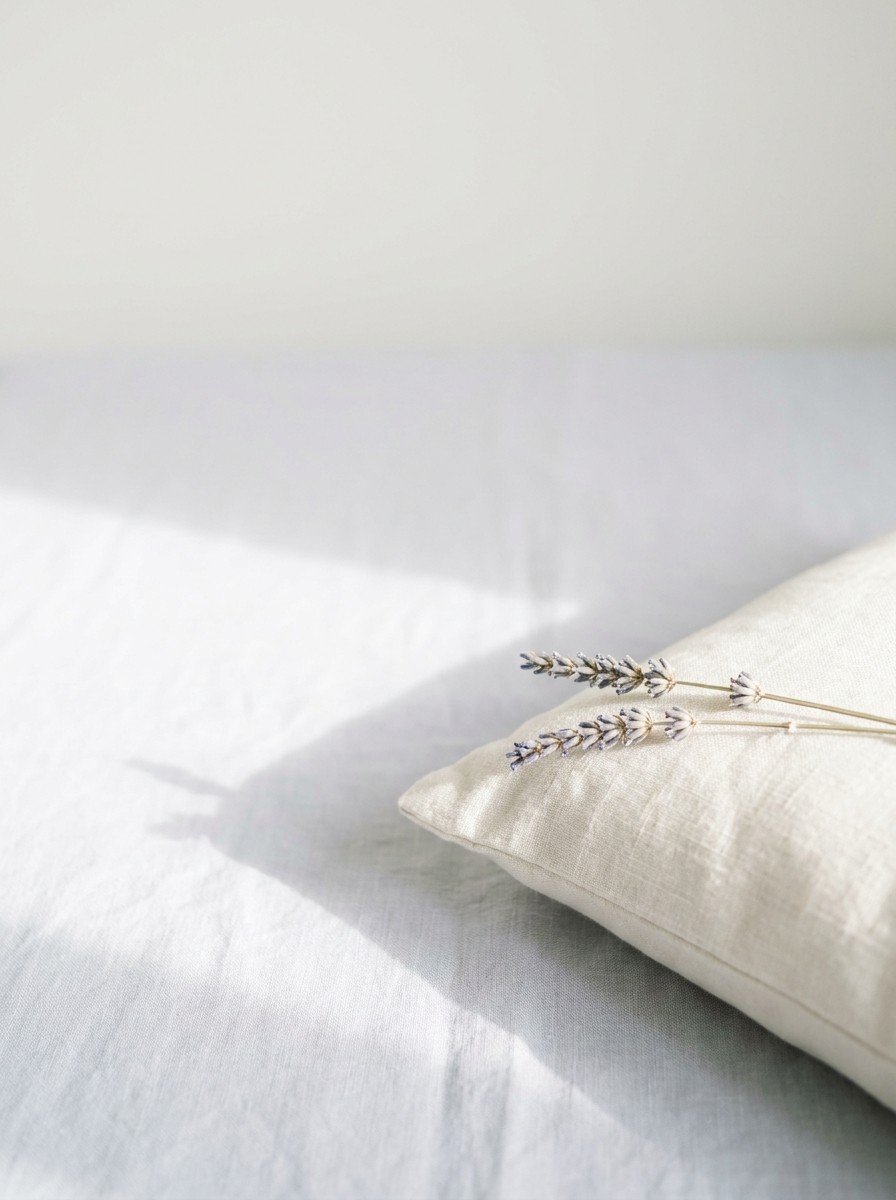 The corner of a linen pillow with two dried lavender stems resting on it in the lower right of pale gray linen, upper left empty