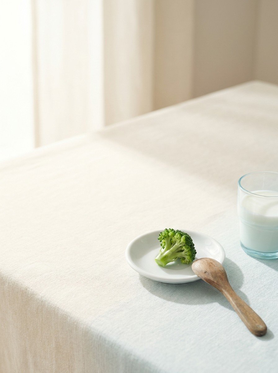 A single steamed broccoli floret on a tiny white saucer, a wooden child's spoon and a glass of milk on cream linen, upper left of the frame left empty