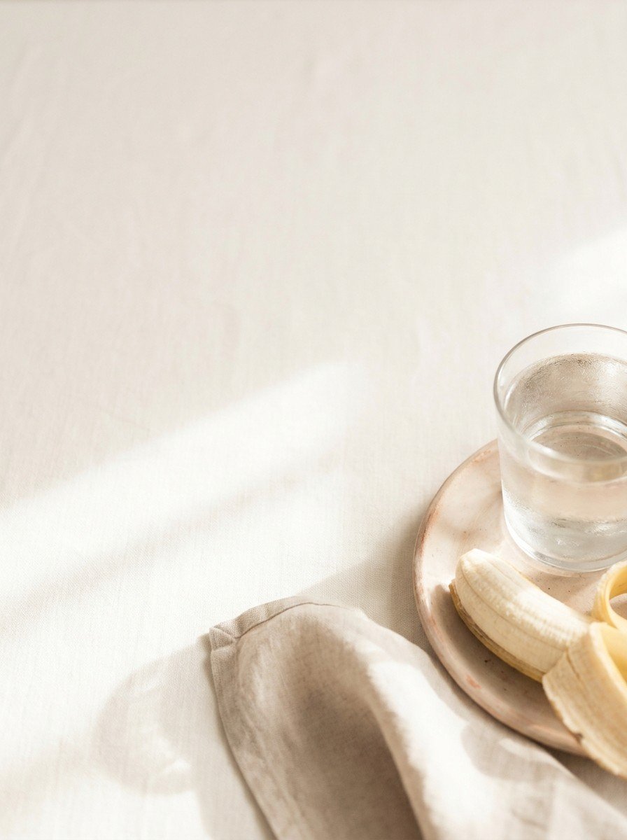 A glass of warm water and half a banana on a clay plate with a linen napkin in the lower right of cream linen, upper left empty