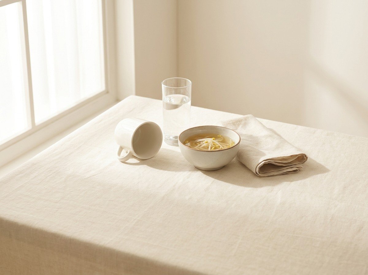 An upturned small white ceramic cup, a tall glass of water, a small bowl of clear bean sprout soup and a folded linen napkin grouped on cream linen in soft window light, with open linen below.