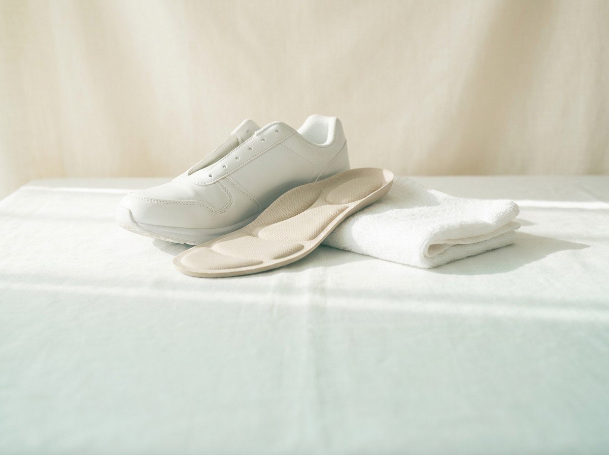 A plain white running shoe without laces, a beige cushioned insole and a folded white towel on white linen in soft window light