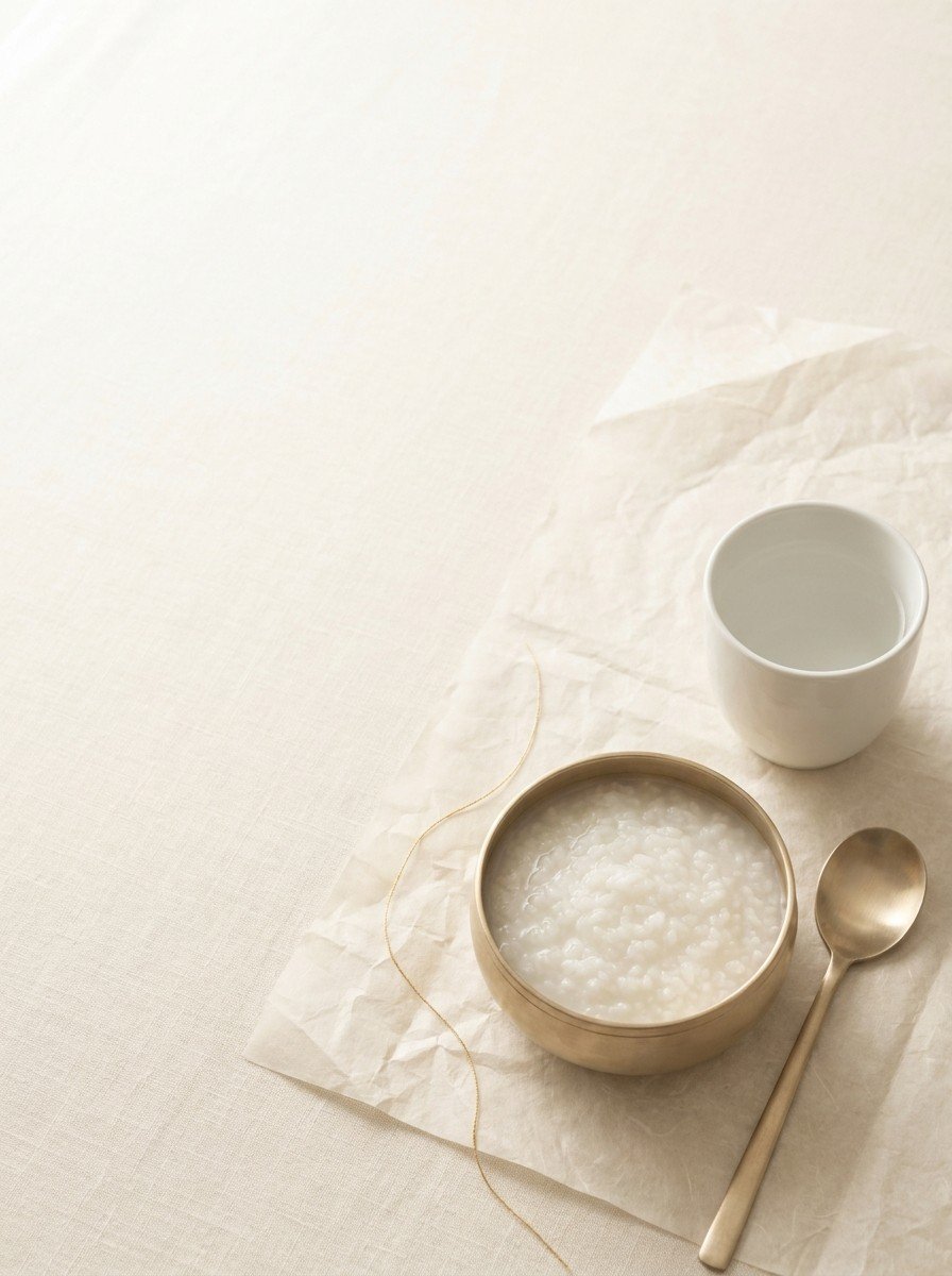 A small brass bowl of plain rice porridge with a brass spoon, a white cup of water and a thin gold thread on cream linen and hanji paper, upper left of the frame left empty