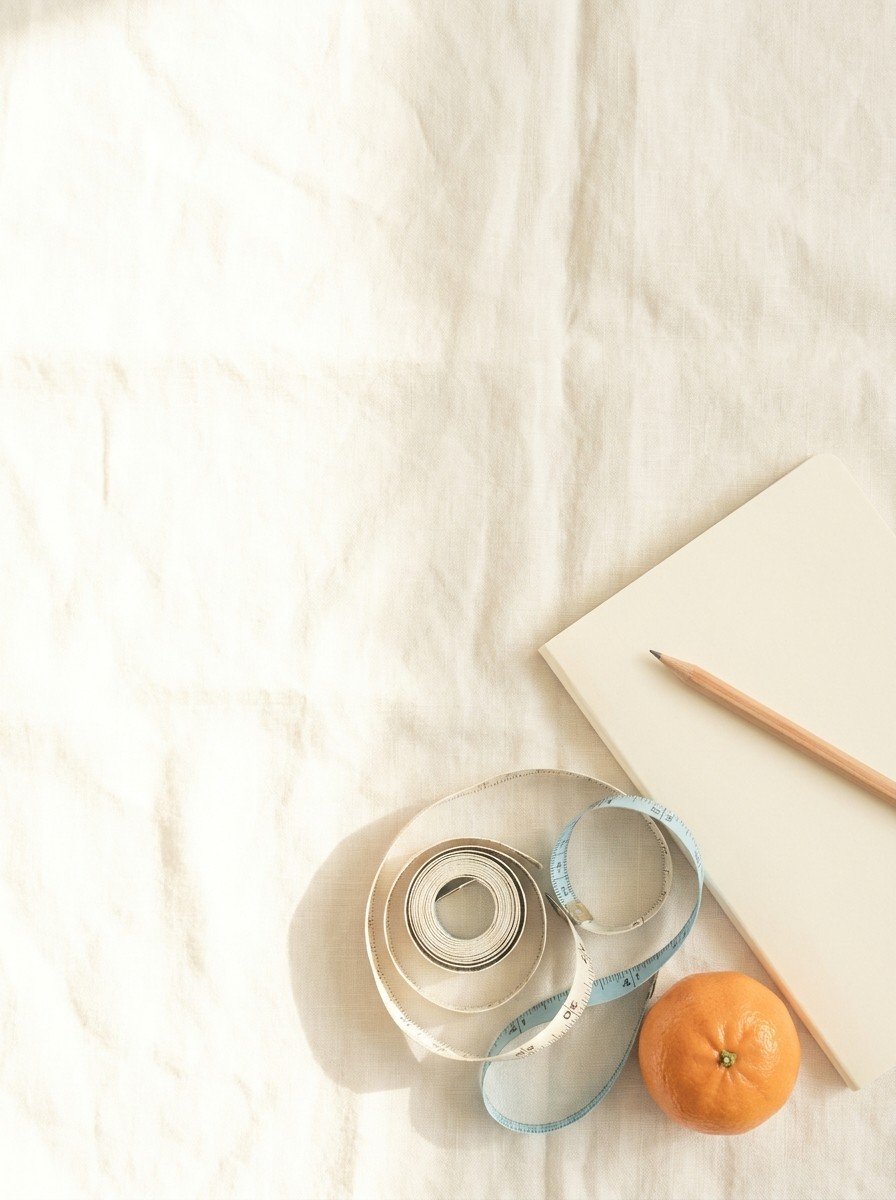 A plain cream notebook with a pencil, a loosely coiled cloth measuring tape and a tangerine on the lower right of cream linen, upper left of the frame left empty