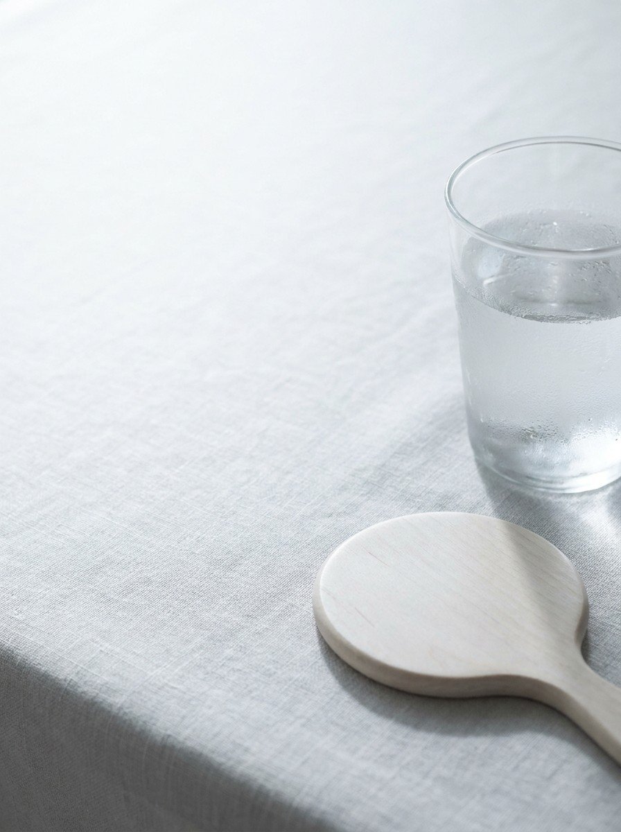 A glass of cold water with droplets and a face-down round hand mirror in the lower right of pale gray linen, upper left empty