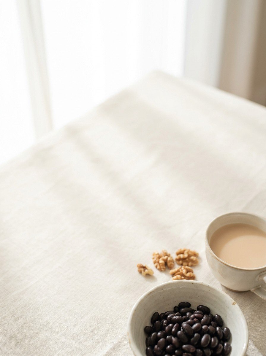 A small bowl of cooked black beans, a few walnuts and a cup of pale tea at the lower-right of a cream linen table, the upper-left left empty in soft window light.