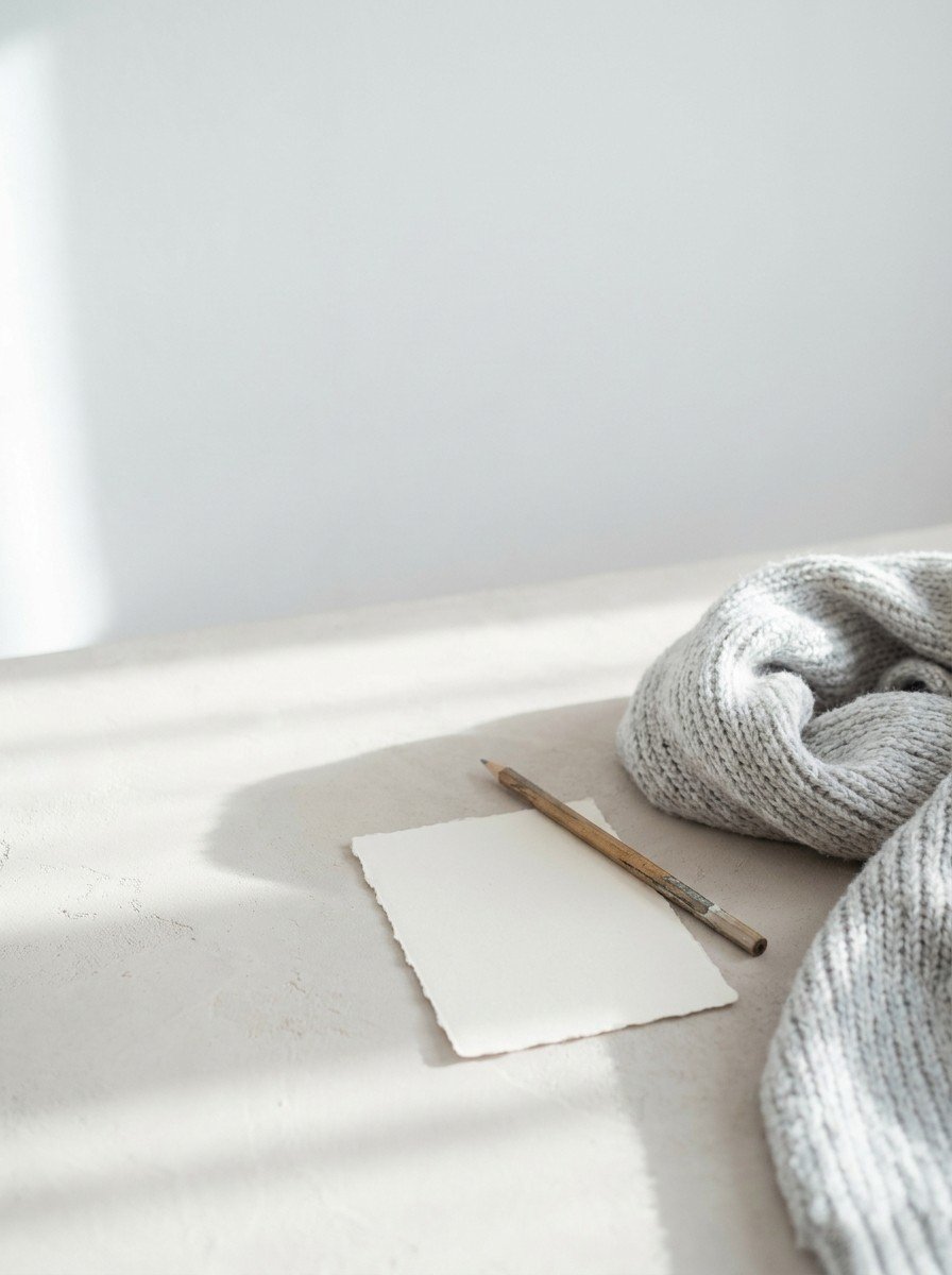 A loosely unwound gray knitted scarf and a blank paper card with a pencil in the lower right of a pale plaster surface, upper left empty