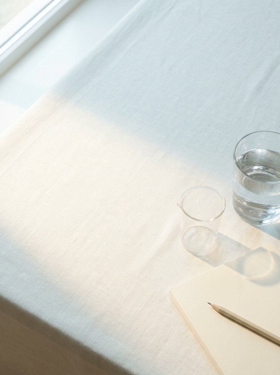 A clear glass of water, a small plain glass measuring cup and a cream notebook with a pencil on the lower right of cream linen, upper left of the frame left empty
