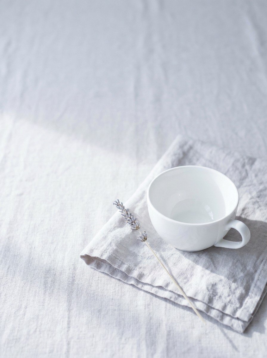 A white porcelain teacup on a folded pale linen napkin with a lavender stem in the lower right of pale gray linen, upper left empty