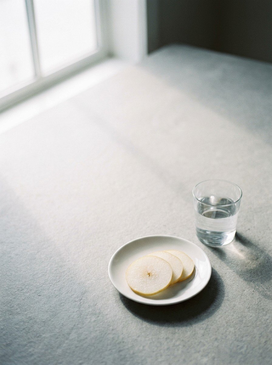A small white plate of sliced Asian pear and a glass of water in the lower right of a light gray stone surface, upper left empty