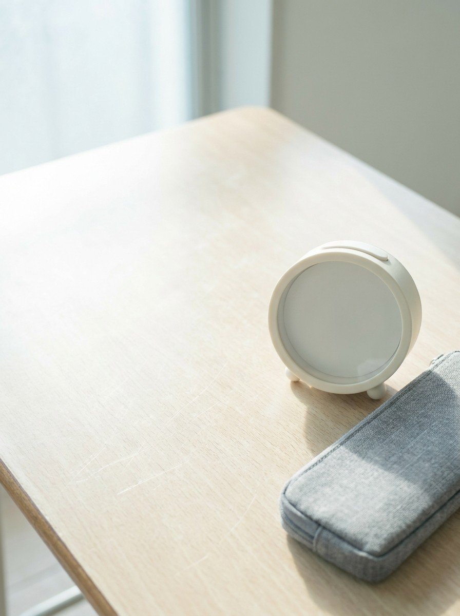 A small round alarm clock with a plain dial and a gray fabric pencil case in the lower right of a pale wooden school desk, upper left empty