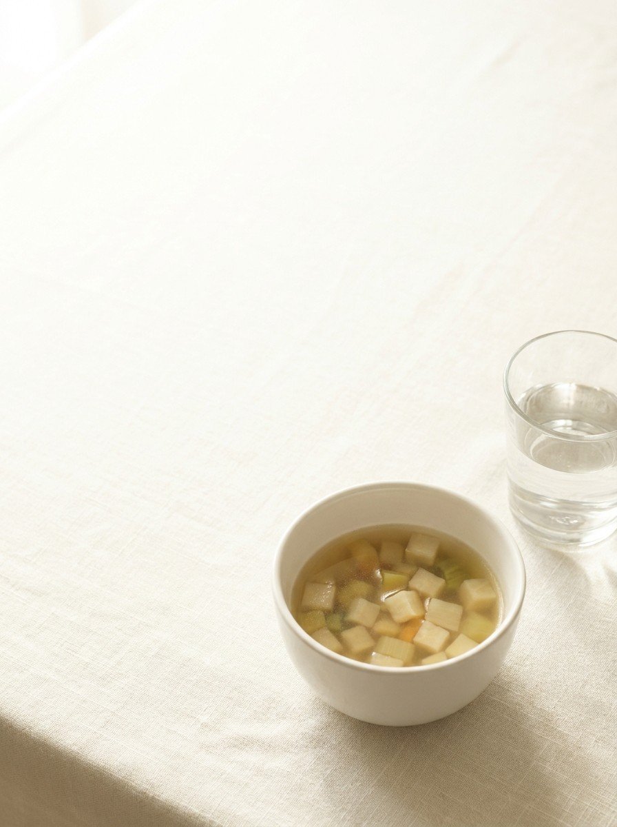 A small ceramic bowl of clear soup with only vegetables and a glass of water at the lower-right of a cream linen table, the upper-left left empty in soft window light.