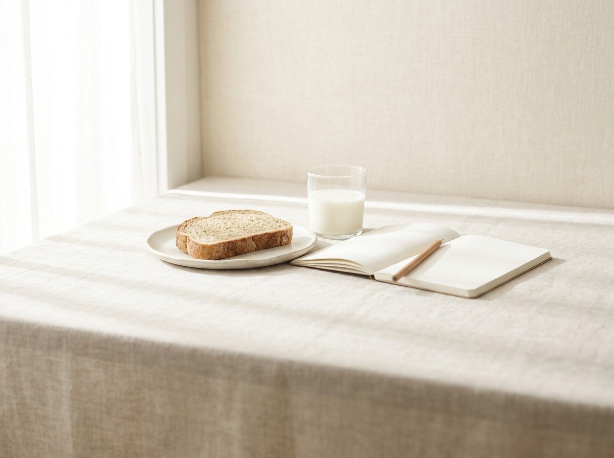 A slice of rustic wheat bread on a small plate, a short glass of milk and an open blank notebook with a pencil clustered on oatmeal linen, lower third empty