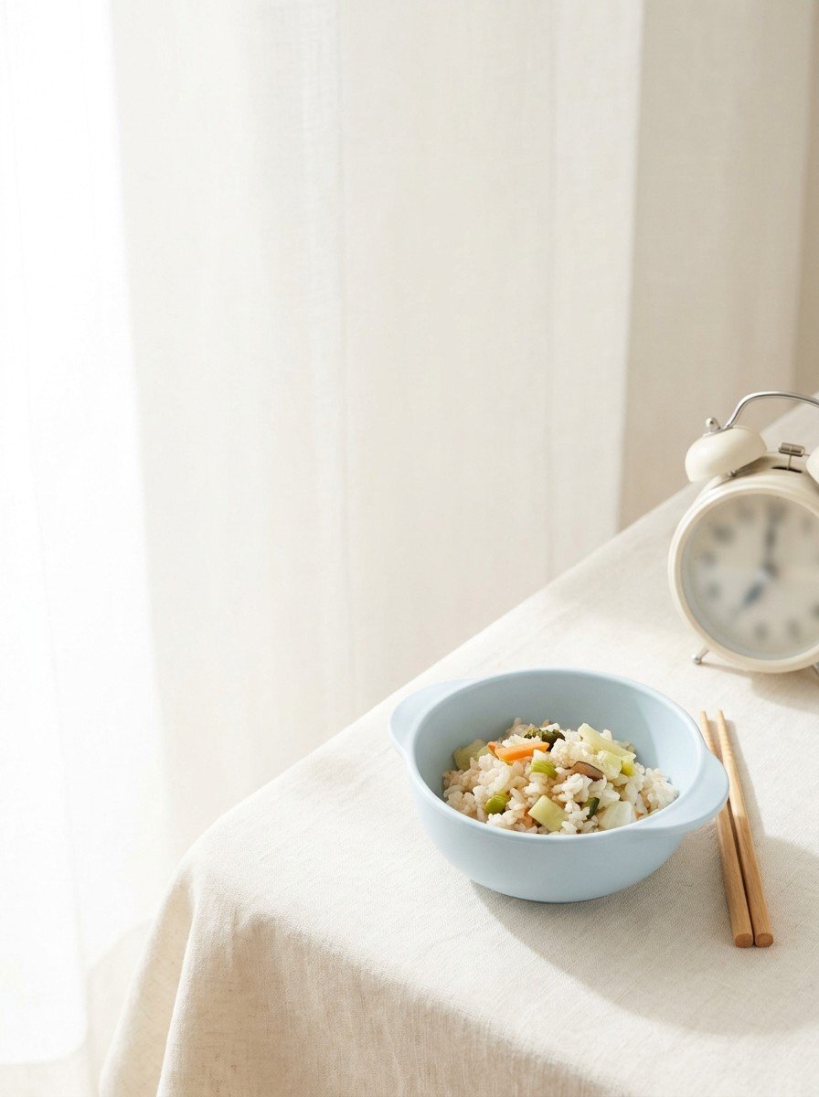 A pale blue child's rice bowl, wooden chopsticks and a cream alarm clock on the lower right of cream linen, upper left of the frame left empty