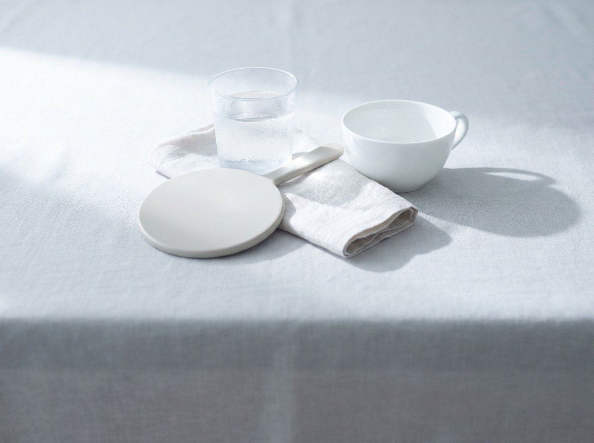 A small round hand mirror lying face-down, a glass of cold water, a folded pale linen napkin and a white teacup clustered in the middle of pale gray linen in window light