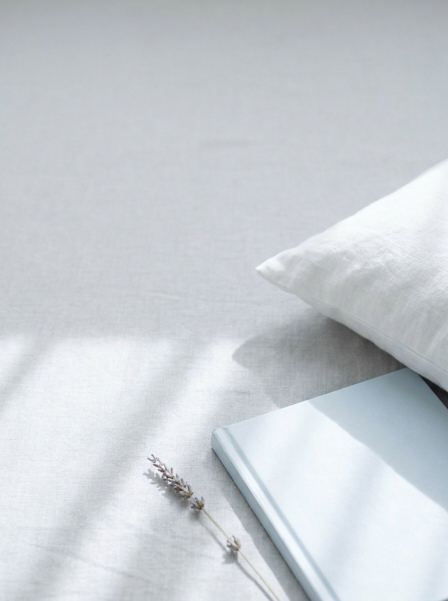 The corner of a white linen pillow, a closed pale-blue picture book and a lavender stem in the lower right of pale gray linen, upper left empty