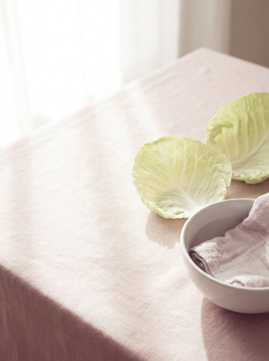 Two fresh cabbage leaves and a small white bowl of cool water with a damp cloth in the lower right of blush linen, upper left empty