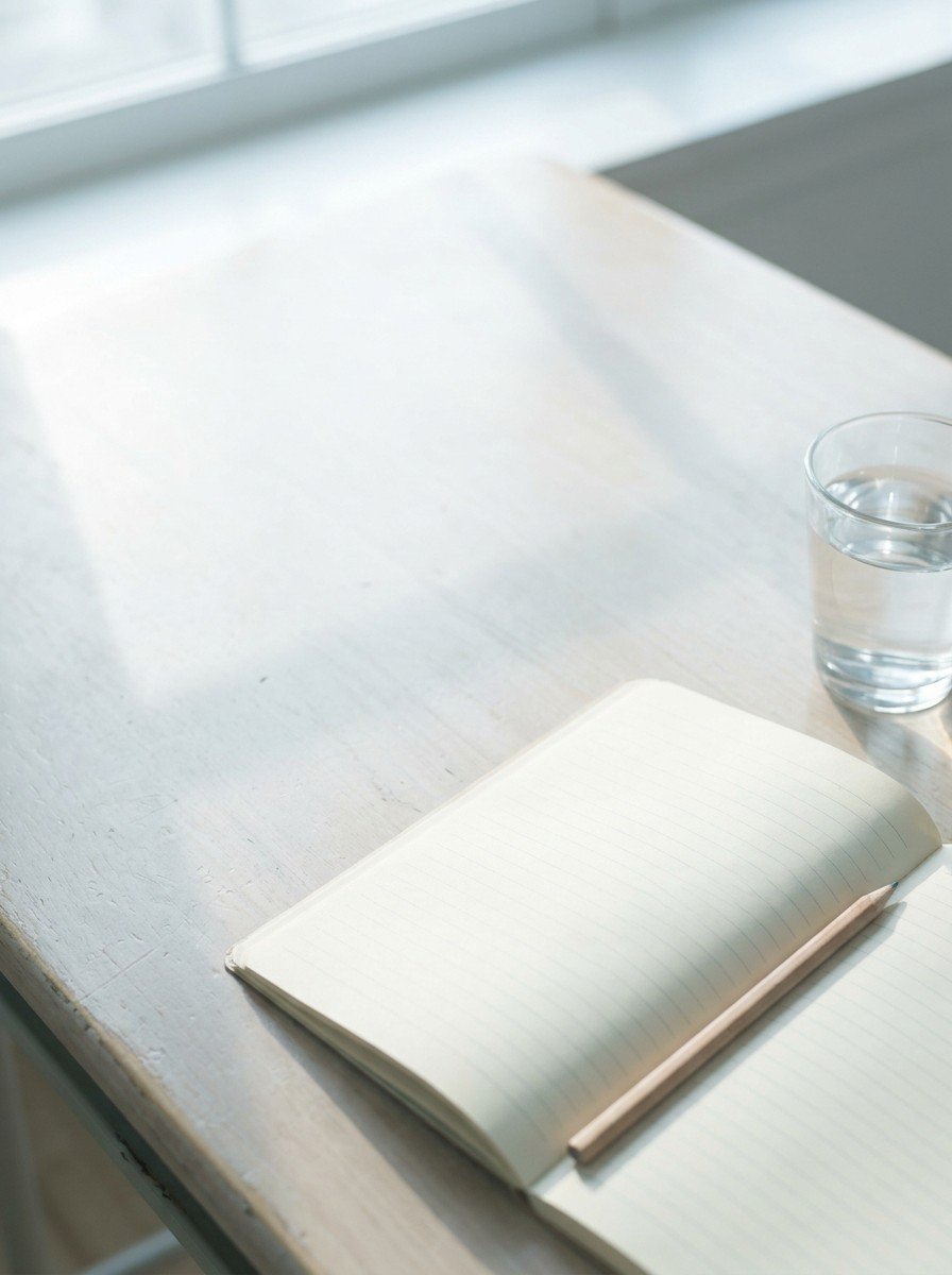 An open lined notebook with blank pages, a pencil and a clear glass of water in the lower right of a pale wooden school desk, upper left empty