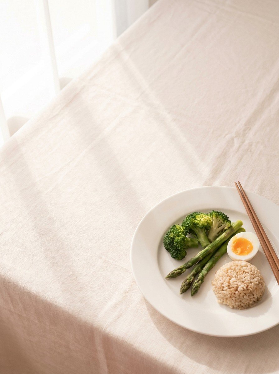 A white plate of steamed green vegetables, a halved boiled egg and a small mound of brown rice with chopsticks at the lower-right of a cream linen table, the upper-left left empty in soft window light.