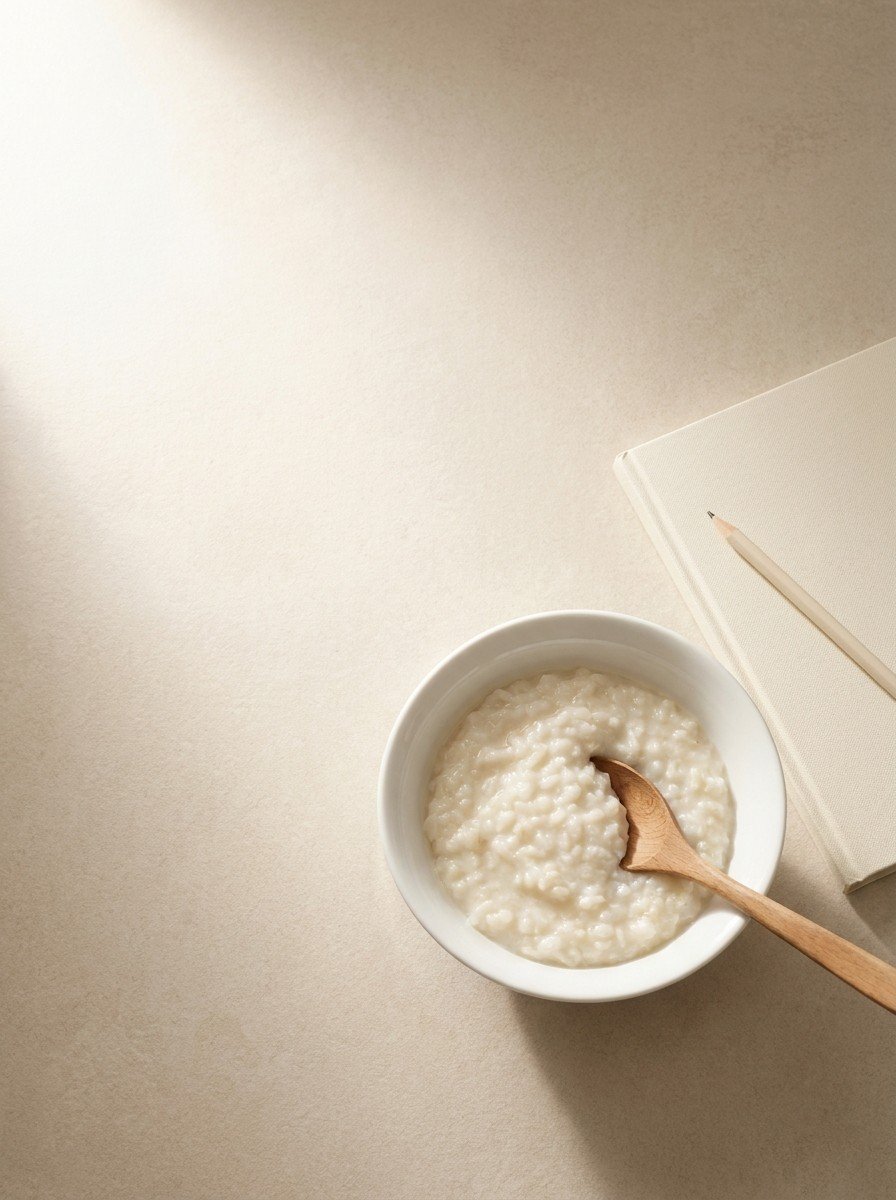 A white porcelain bowl of rice porridge with a wooden spoon and a closed notebook with a pencil in the lower right of pale stone, upper left empty