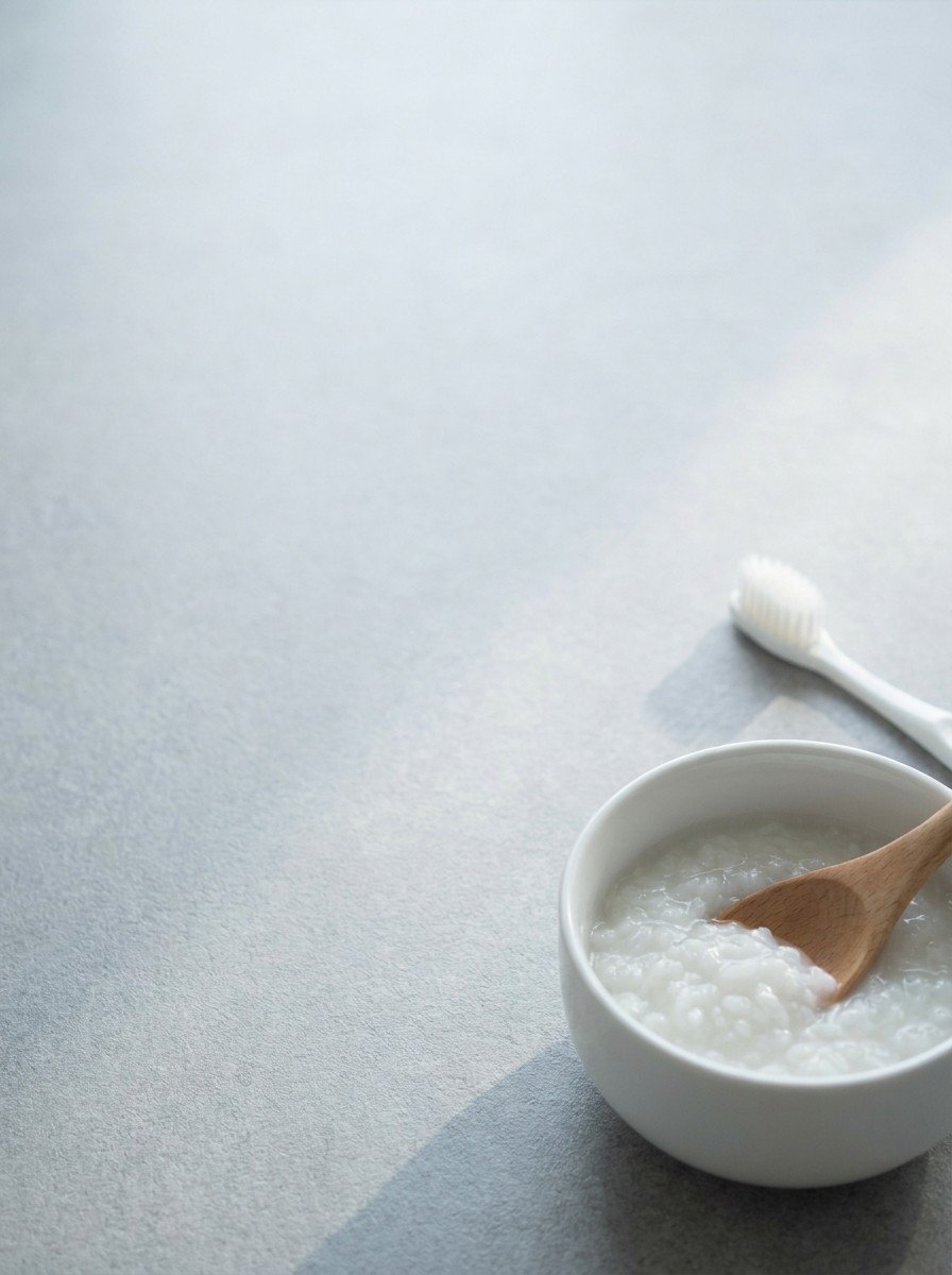 A small white bowl of plain rice porridge with a wooden spoon and a white toothbrush in the lower right of a light gray stone surface, upper left empty